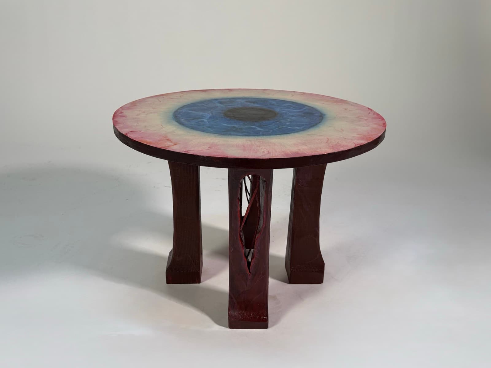Handmade round table with a painted blue eye across the tabletop and dark reddish wooden legs, three-quarter view