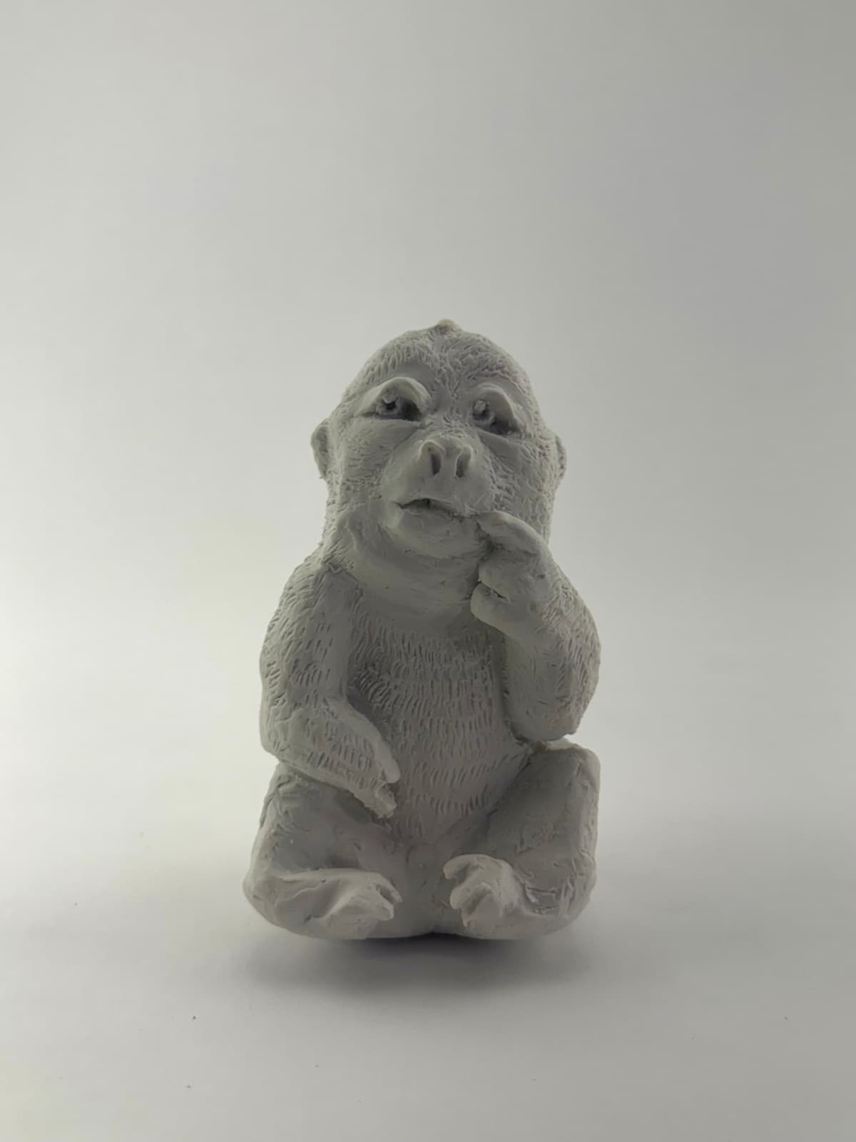 Small light-colored hand-sculpted monkey seated with one finger raised near its mouth, front view