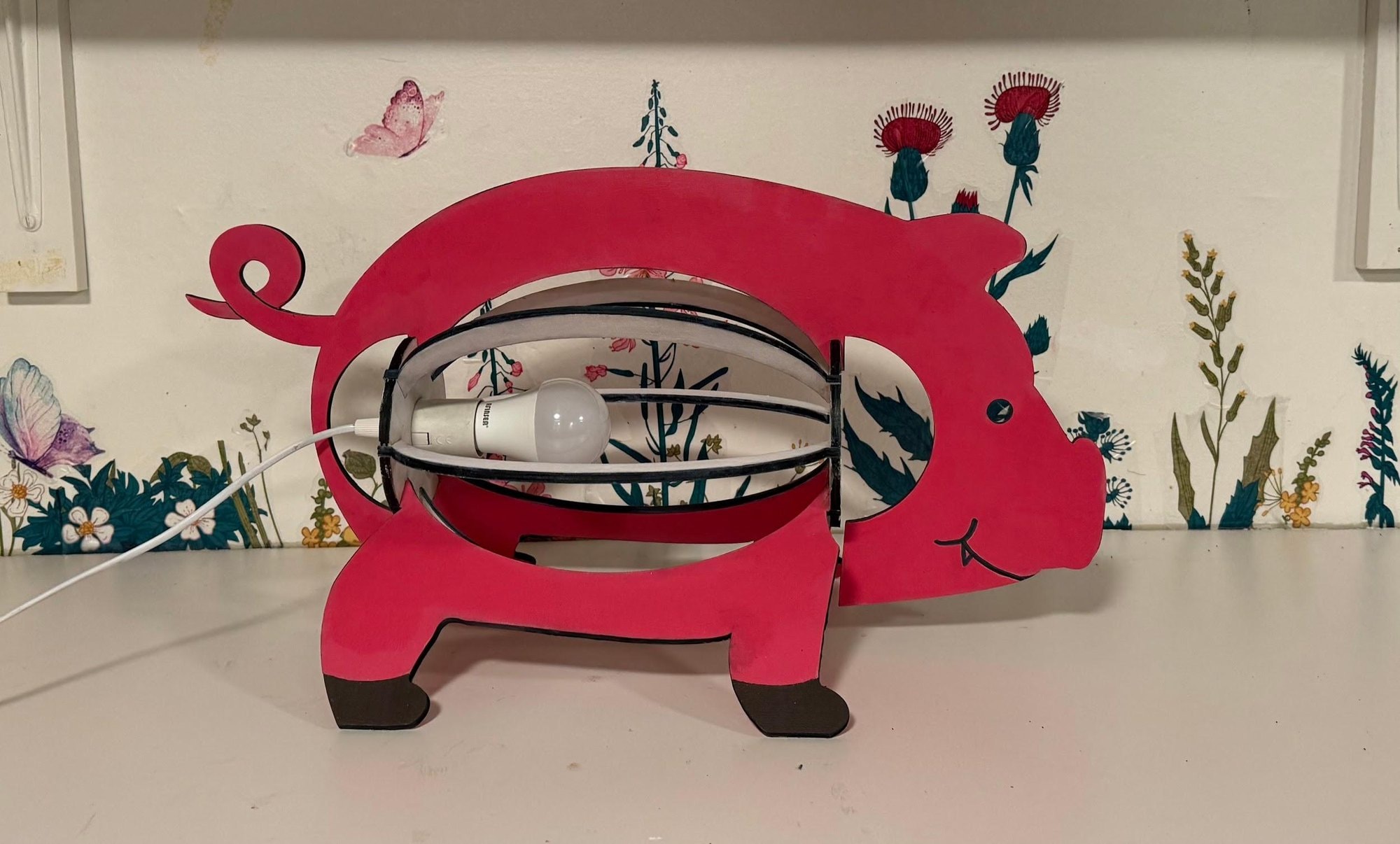 The finished pink pig lamp on a table in front of a wall painted with flowers and butterflies.