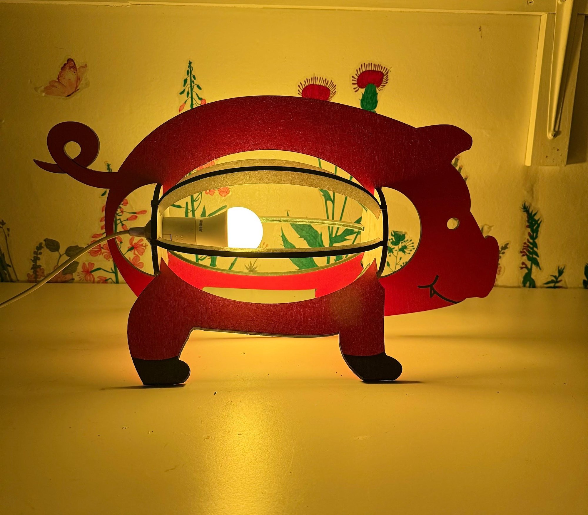 The pink pig lamp switched on in a dim room, glowing warm yellow through the cage of ribs that forms its body.